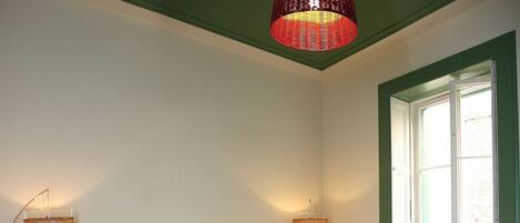 A bedroom with a large bed, two bedside lamps, a window with green shutters, and a hanging red pendant light.