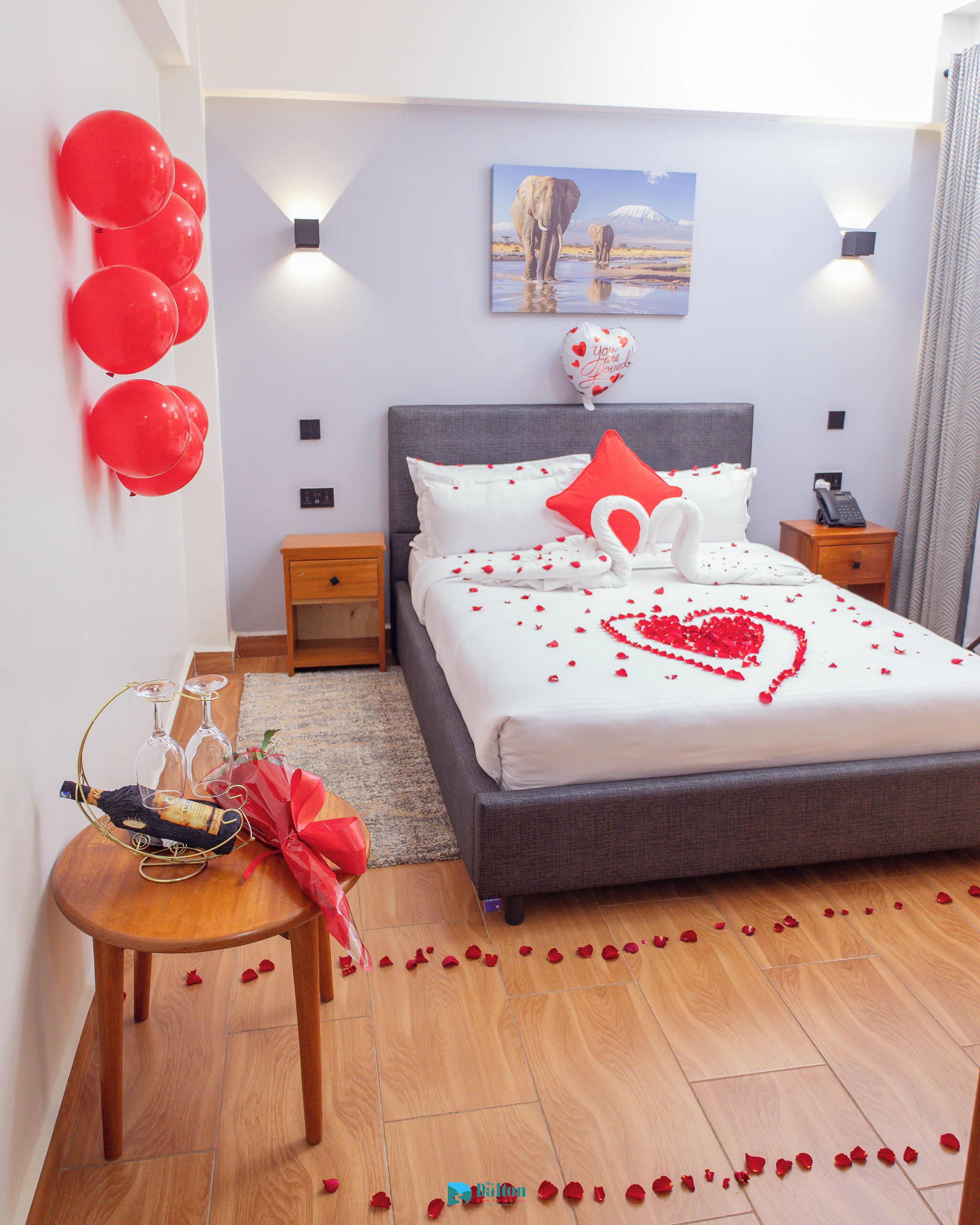 A bedroom with a bed, a bedside table with a bottle and glasses, a nightstand with a phone, and a wall with a picture and balloons.