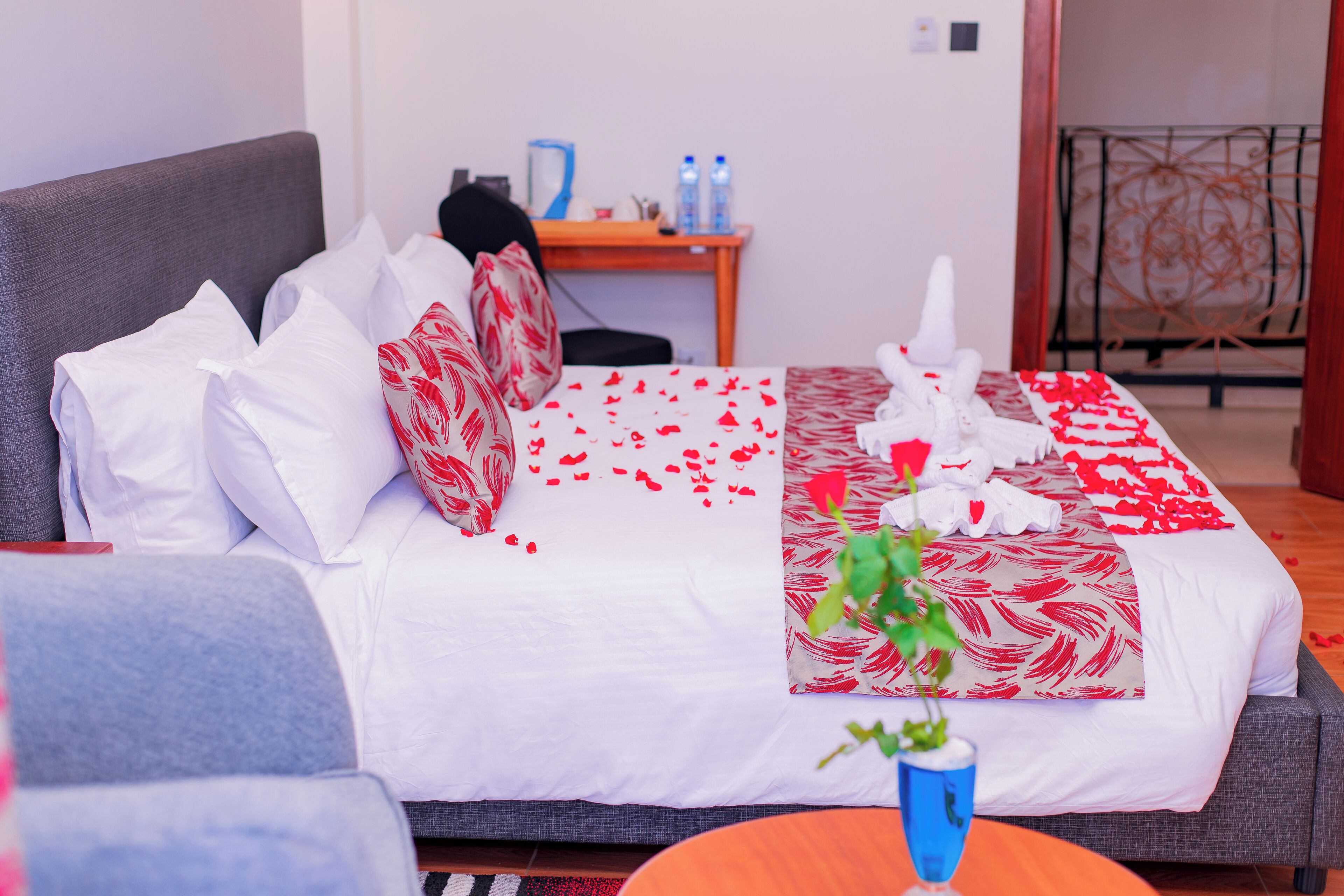 Una cama bien hecha con ropa blanca y roja, una mesita con un florero azul y un sofá gris.