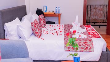 Una cama bien hecha con ropa blanca y roja, una mesita con un florero azul y un sofá gris.