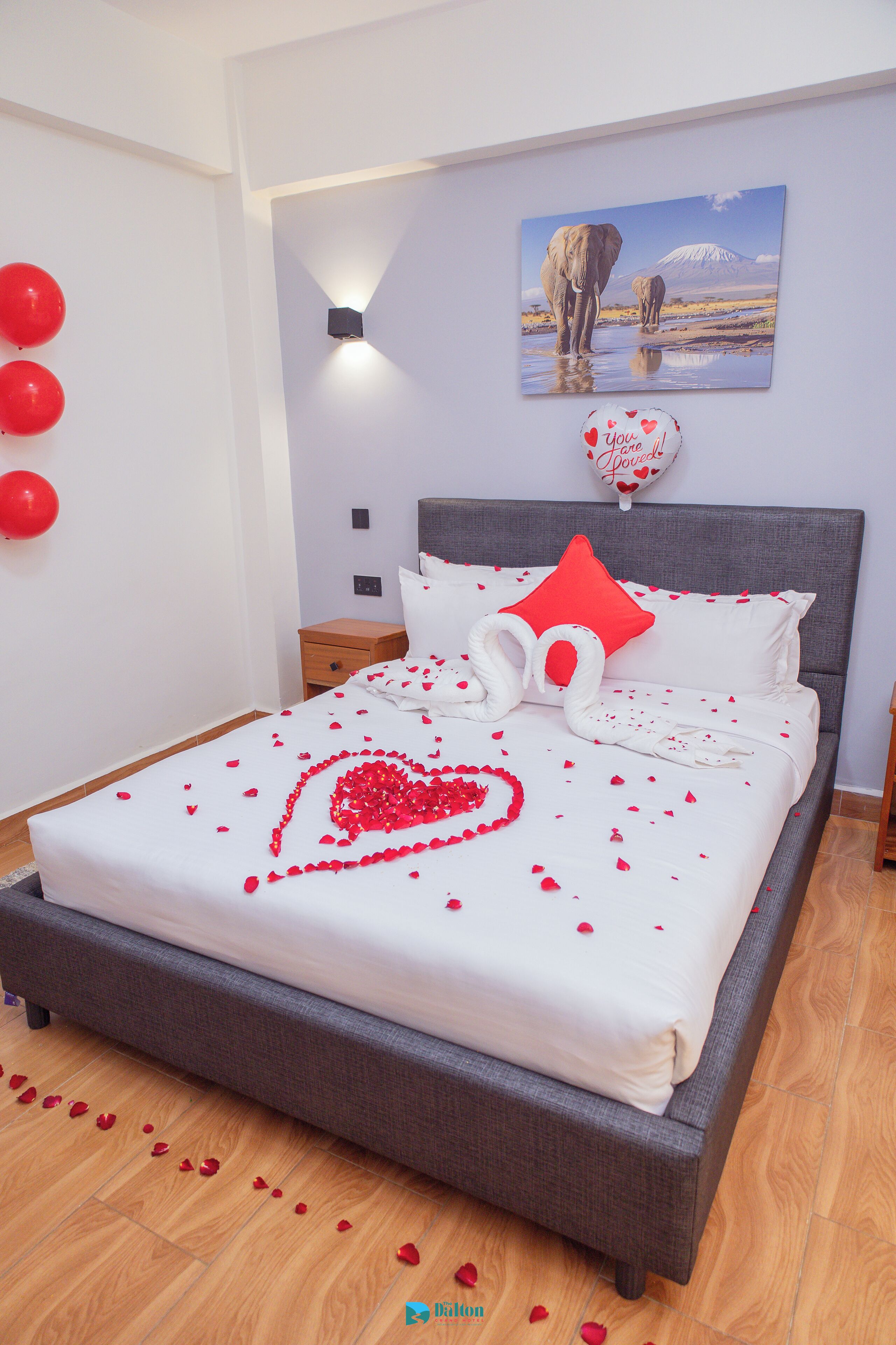 A neatly made bed with rose petals and a heart design, a wall with a picture of elephants and a mountain, and red balloons on the wall.