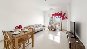 A modern living room with a dining area, a sofa, a TV, and a large window with a view of buildings. - FIRST CLASS | 2BR | Cozy City Retreat (JLT)