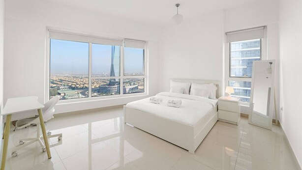 A modern, minimalist bedroom with a large bed, a desk, and a view of a cityscape through large windows. - FIRST CLASS | 2BR | Cozy City Retreat (JLT)
