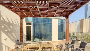 A covered outdoor seating area with a wooden table and chairs, a glass door leading inside, and a view of a modern building. - FIRST CLASS | 2BR | Cozy City Retreat (JLT)