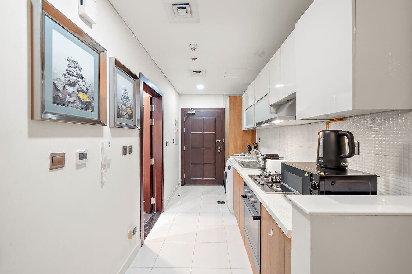 A narrow hallway leading to a kitchen with white cabinets, a stove, and a kettle.