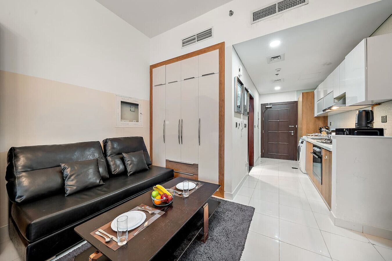 A modern apartment with a black leather sofa, a dining table set with plates and a fruit bowl, a kitchen with a stove and refrigerator, and a wardrobe with sliding doors.