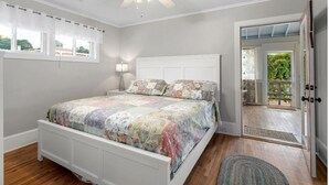 Een slaapkamer met een wit bed, een plafondventilator, een raam met gordijnen en uitzicht op de omgeving door een open deur.