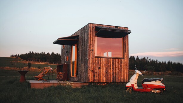 Une cabane en bois rustique dotée d’une grande fenêtre, d’une terrasse avec des meubles d’extérieur et d’un scooter rouge garé devant.