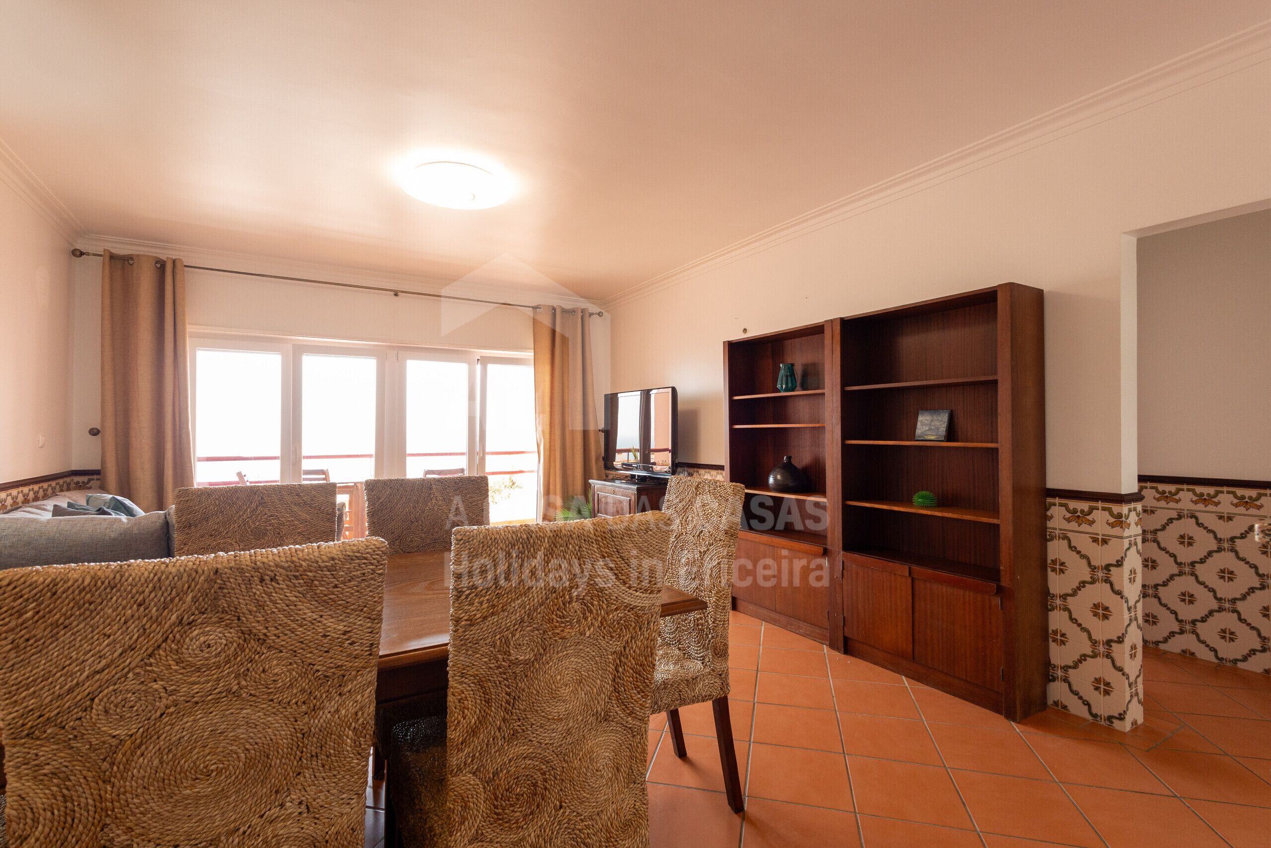 A dining area with a wooden table and chairs, a bookshelf, and a view through a window.