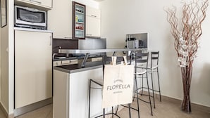 Une cuisine compacte équipée d’un four à micro-ondes, d’un four et de placards de rangement. Un coin bar est aménagé avec une machine à café et un espace détente avec des chaises. Un vase décoratif contenant des fleurs blanches est posé sur le sol.