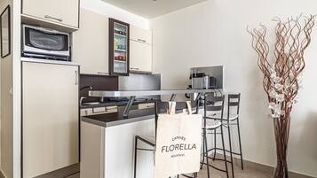 Una cucina compatta con forno a microonde, macchina da caffè e un pensile con stoviglie. Sono presenti due sgabelli da bar e un vaso con fiori bianchi.