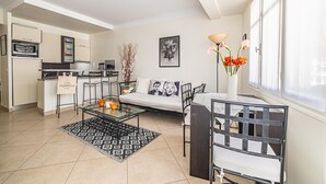 Un salon moderne avec une table basse en verre, un canapé et un coin repas avec une chaise. On aperçoit au fond une cuisine équipée d’un four à micro-ondes et d’un réfrigérateur.
