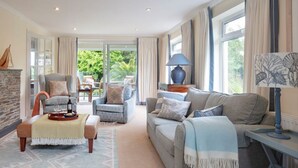 A spacious living room with a fireplace, comfortable seating, and large windows offering a view of greenery. - Luxury, spacious holiday home in St Mawes (St Mawes)