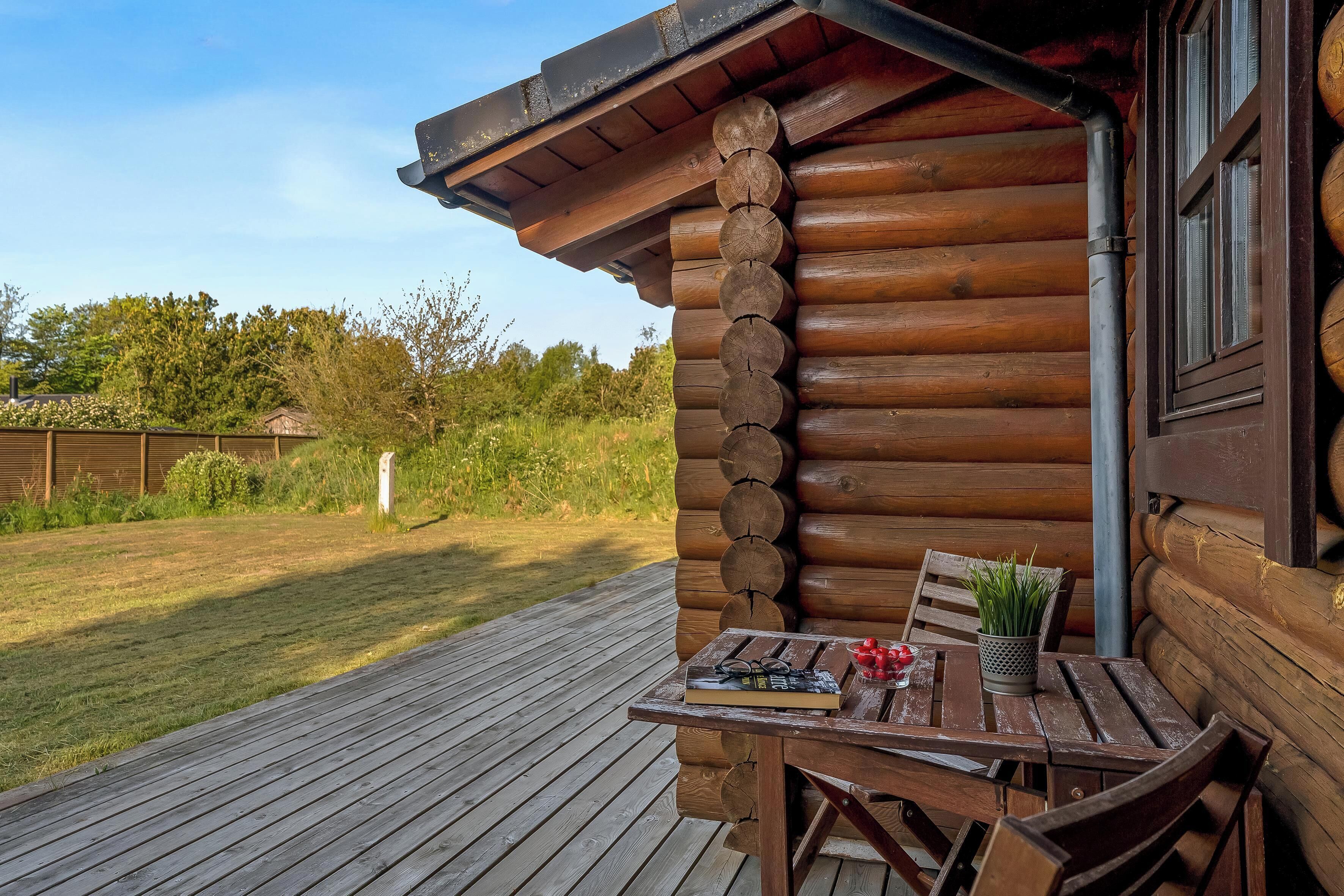 A wooden cabin with a deck, a table with a book and a plant, and a window with a view of the surrounding area.