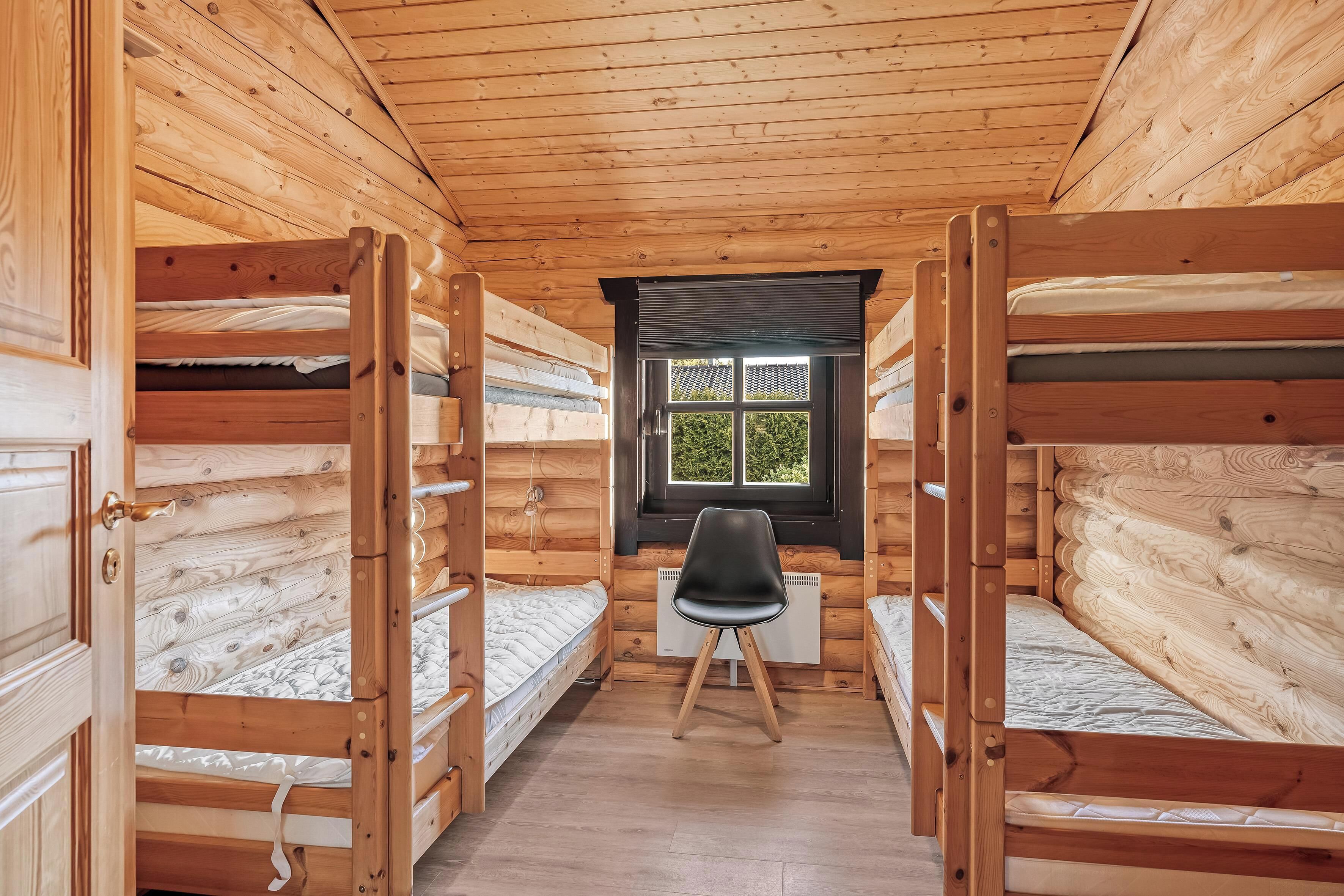 A cabin with wooden walls and floors, bunk beds, a desk, and a window with a view of greenery.