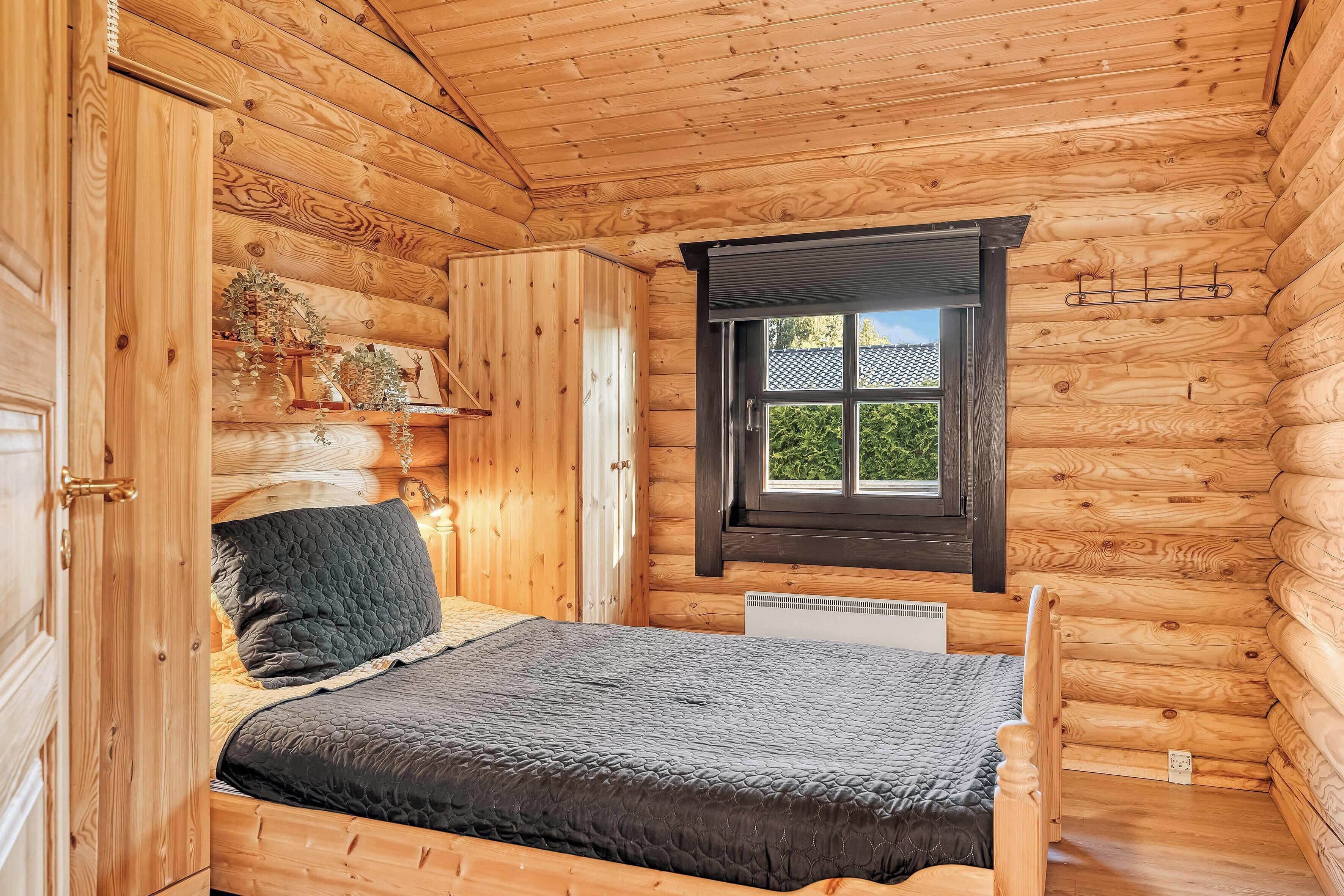 A wooden cabin room with a bed, a window, and a shelf with plants.