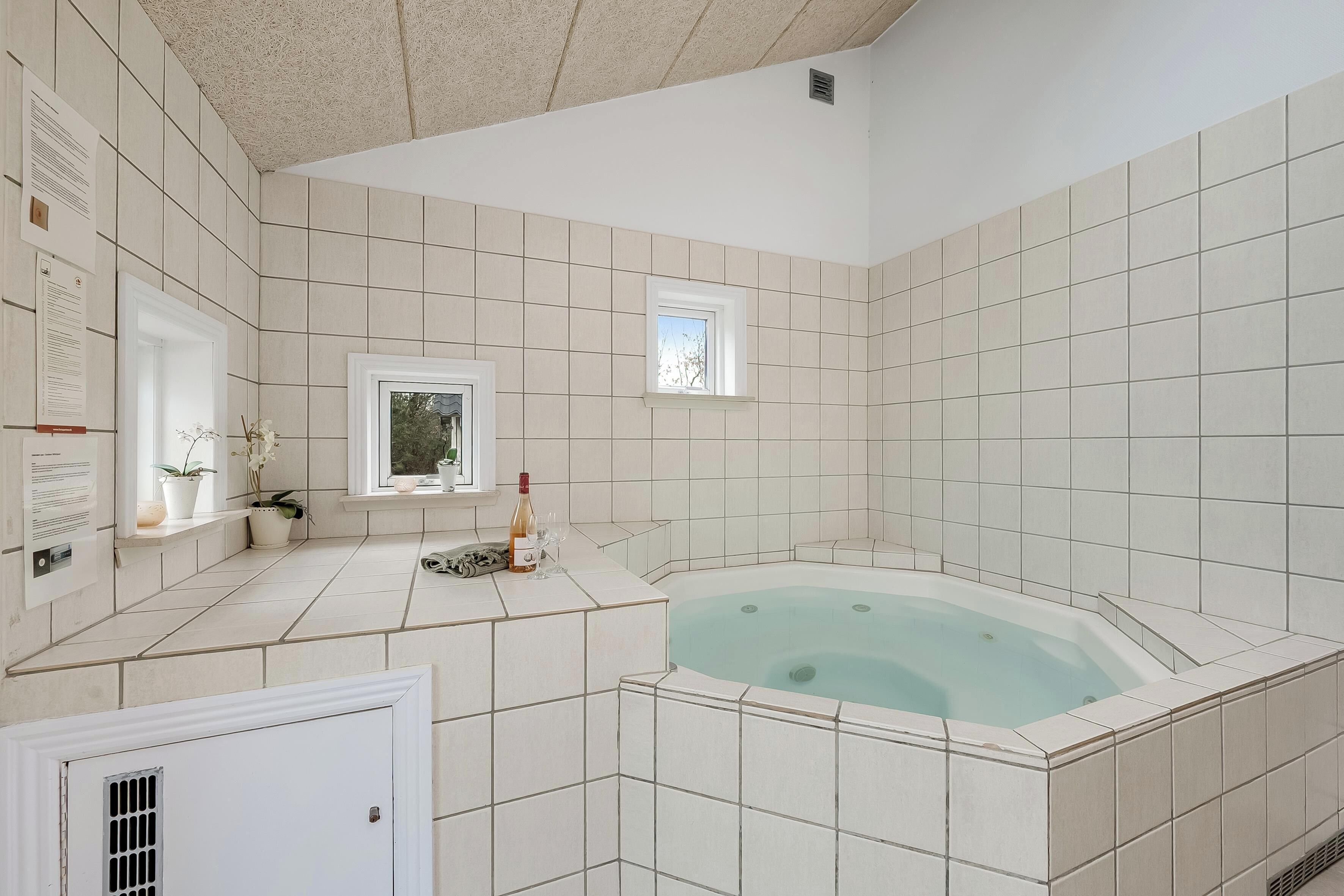 A modern bathroom with a large bathtub, tiled walls, a window, and a small shelf with plants and a bottle.