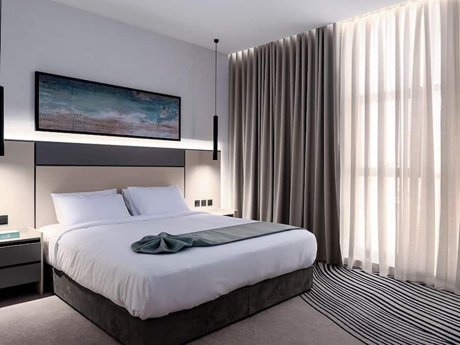 A modern bedroom with a large bed, a nightstand, a wall-mounted TV, and sheer curtains.