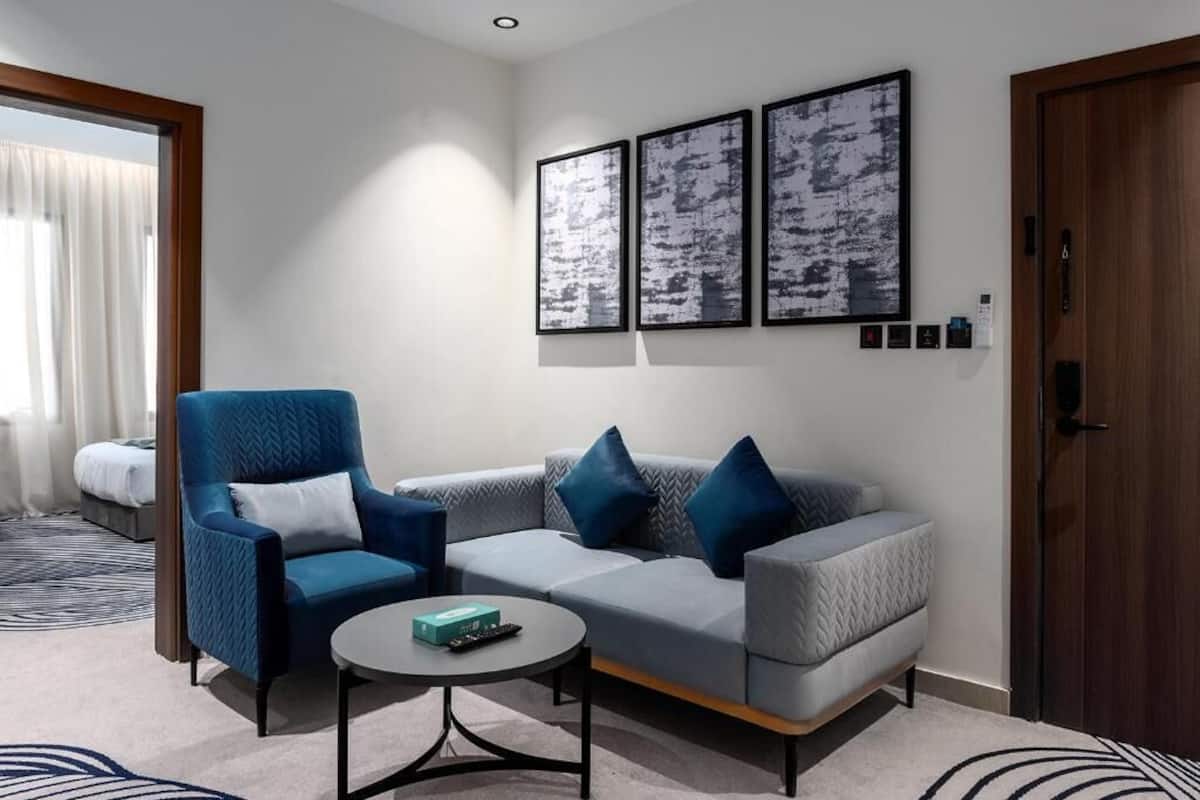 A modern living room with a blue armchair, grey sofa, and a small round table. There are three framed artworks on the wall and a wooden door to the right.