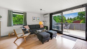 Una sala de estar moderna con un sofá, otomanas, una mesa de centro y una silla. Hay un ventanal corredero que da a un patio al aire libre con una mesa y sillas.