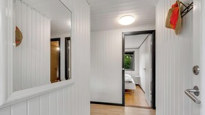 Un pasillo con paredes blancas con paneles, suelo de madera y una puerta que da a un dormitorio con cama y ventana.