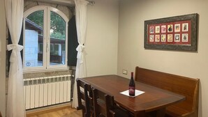 A dining area with a wooden table and chairs, a bottle of wine, a framed picture on the wall, and a window with curtains.
