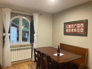 A dining area with a wooden table and chairs, a bottle of wine, a framed picture on the wall, and a window with curtains.