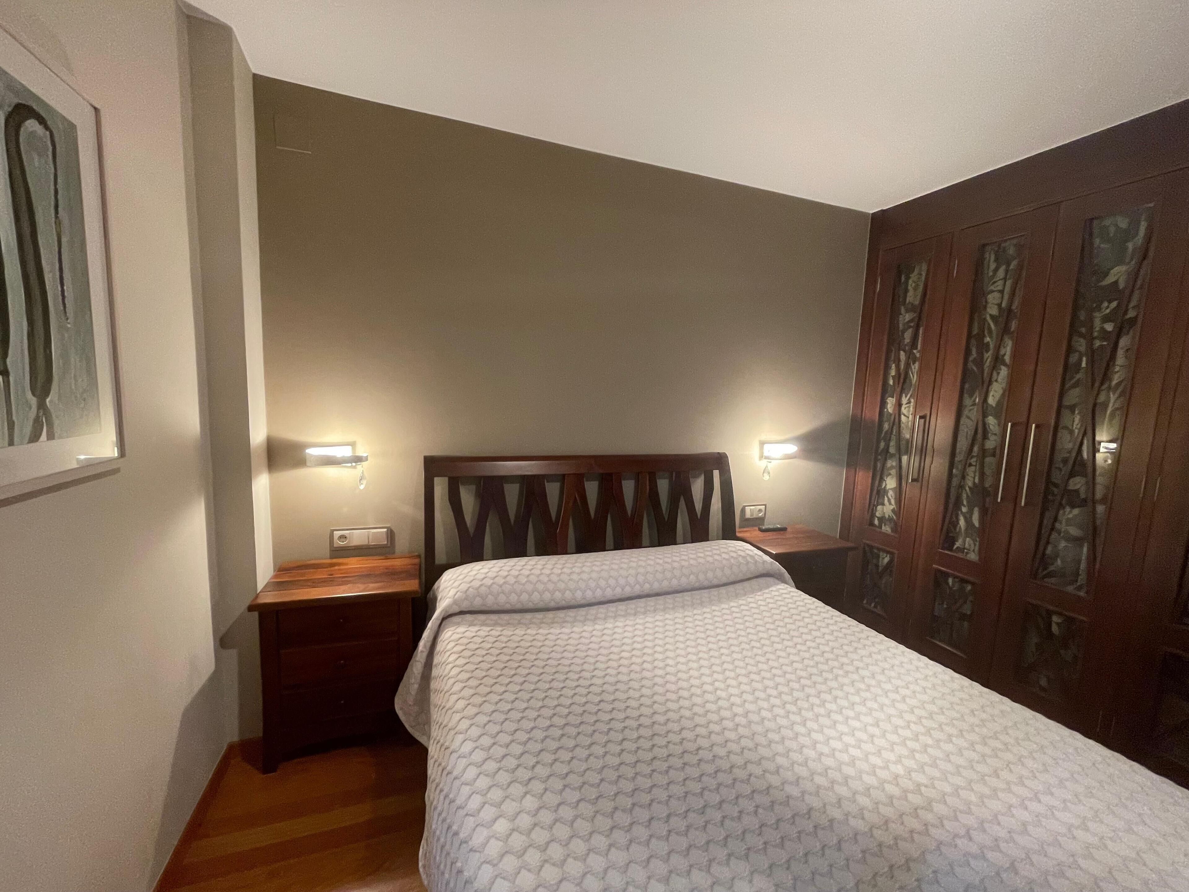 A bedroom with a bed, wooden headboard, two bedside tables with lamps, a nightstand, and a wardrobe with glass doors.