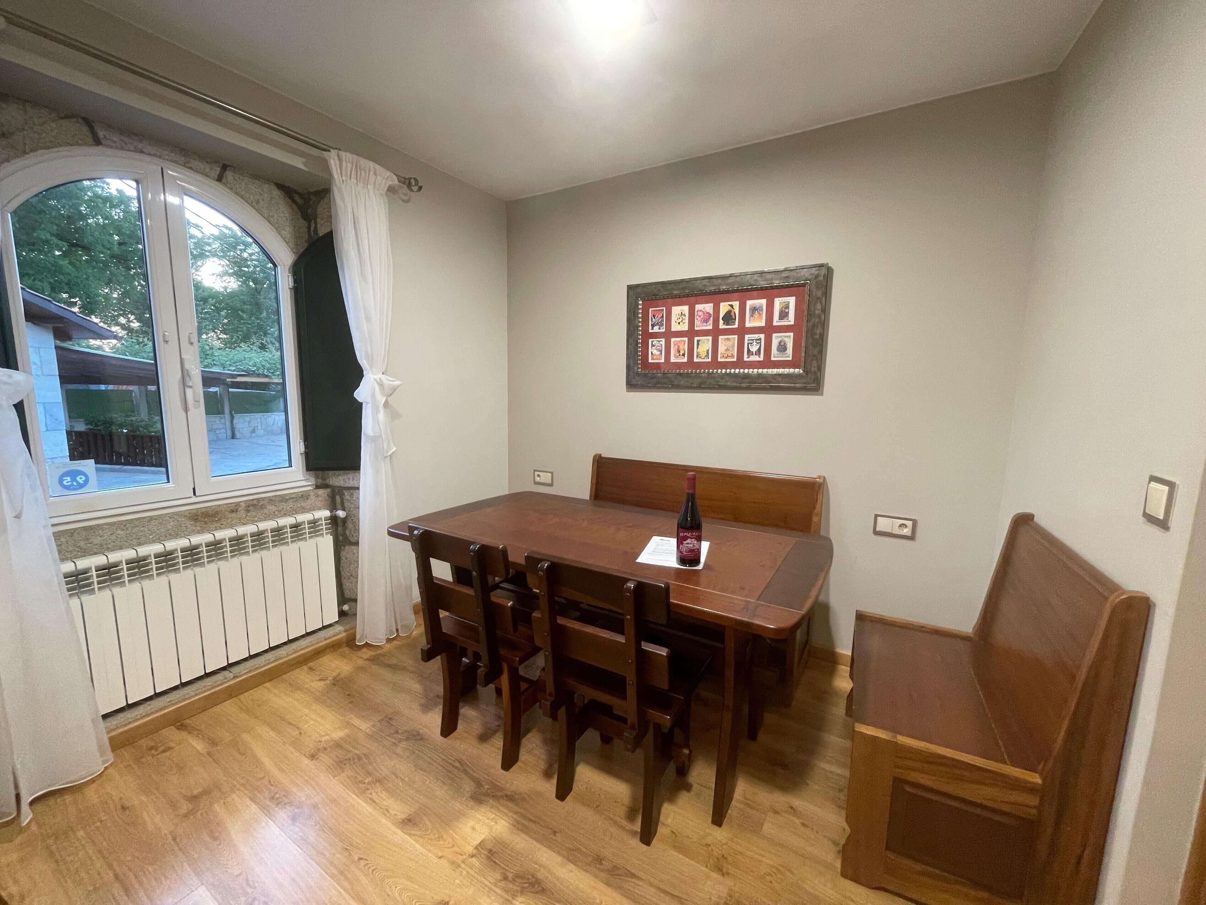 A dining area with a wooden table and chairs, a radiator below the window, and a framed artwork on the wall.