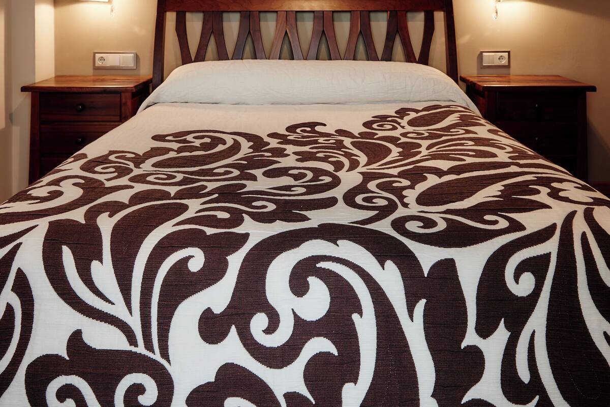 Una cama bien hecha con una colcha estampada, dos mesitas de noche y luces fijadas a la pared.