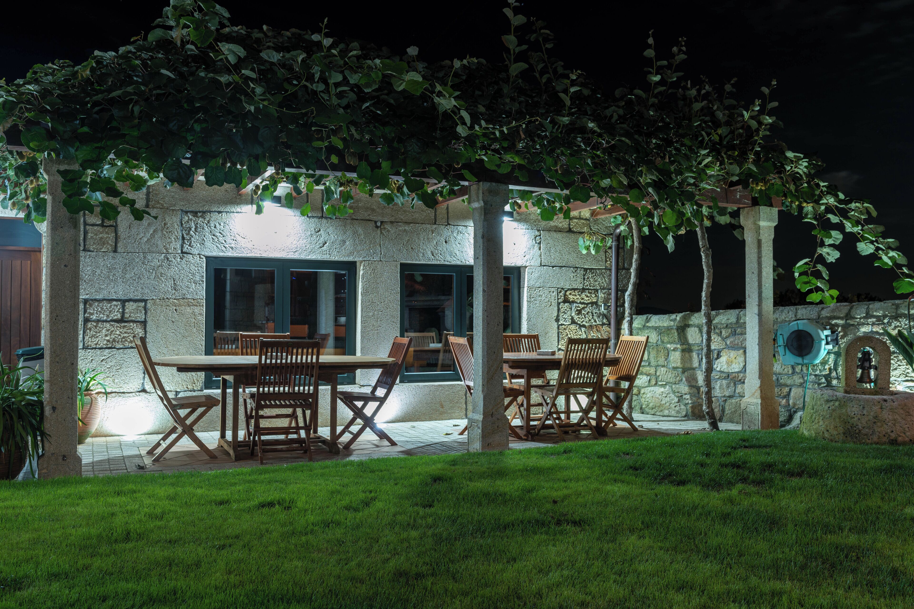An outdoor patio area with stone walls, wooden tables and chairs, and a pergola covered with foliage.