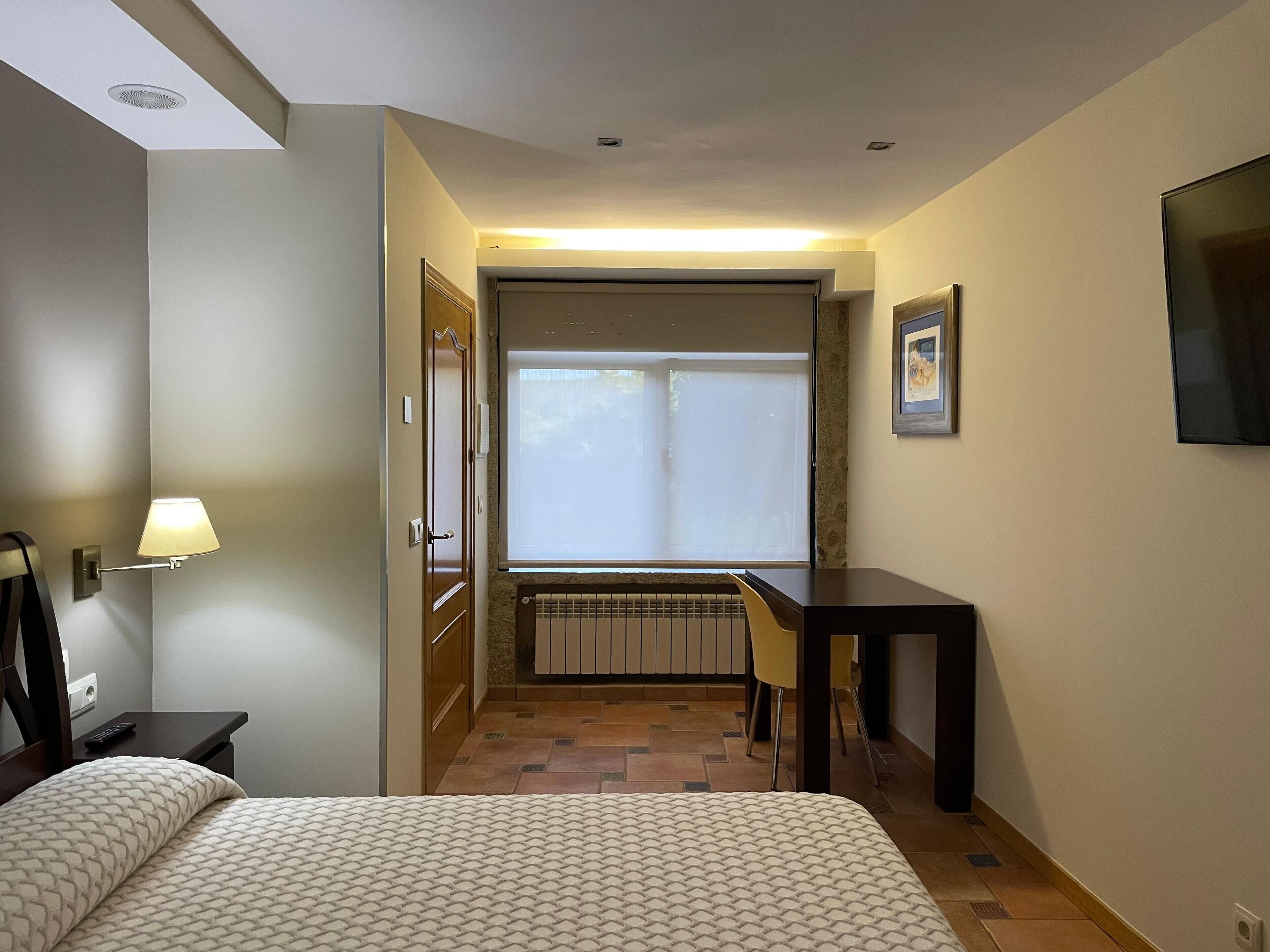A hotel room with a bed, a desk, a chair, a window with blinds, a wall-mounted TV, and a radiator.