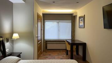 A hotel room with a bed, a desk, a chair, a window with blinds, a wall-mounted TV, and a radiator.