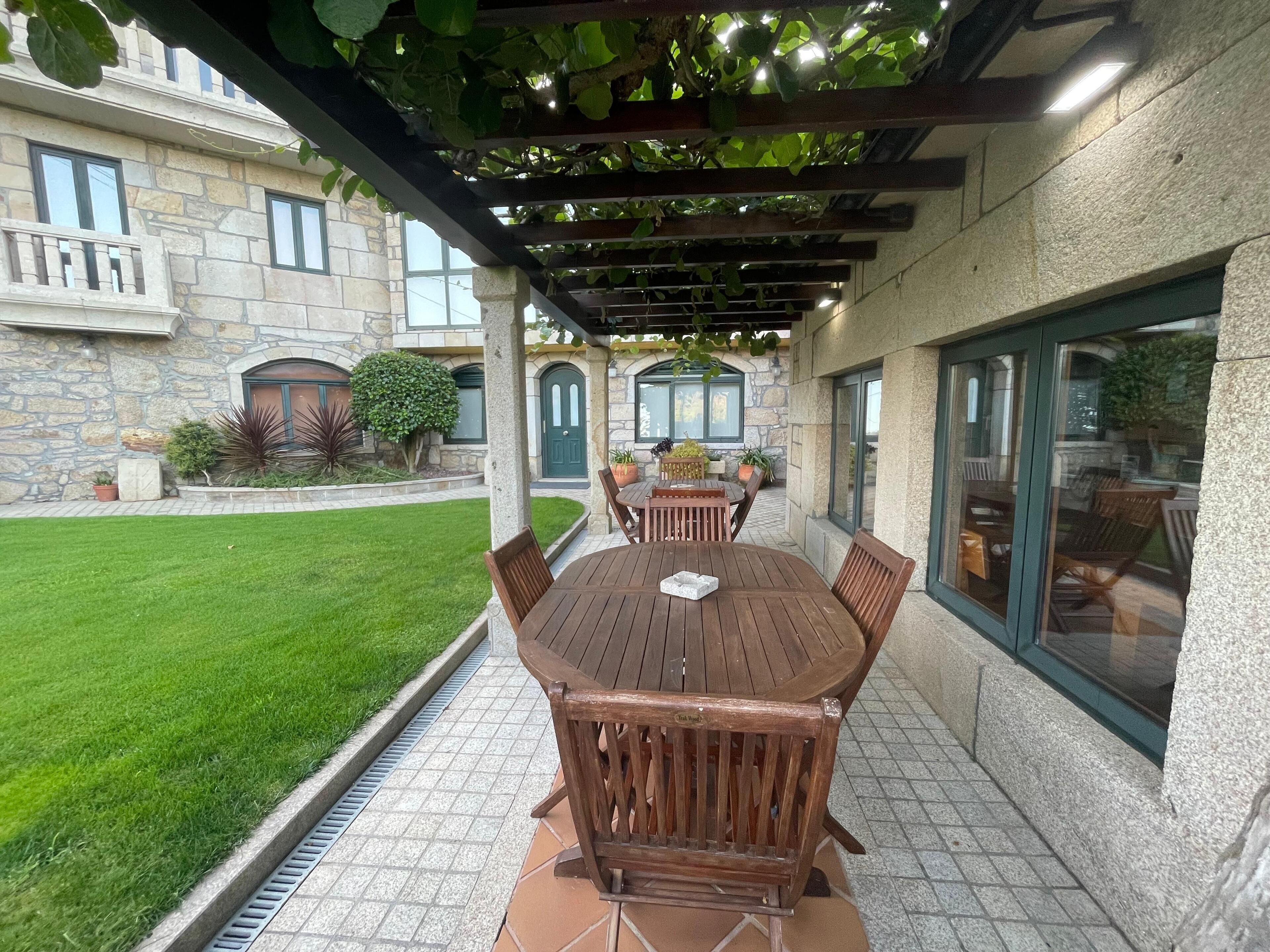 Une terrasse couverte avec une table et des chaises en bois, un bâtiment en pierre en arrière-plan et une pelouse bien entretenue.