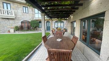 Une terrasse couverte avec une table et des chaises en bois, un bâtiment en pierre en arrière-plan et une pelouse bien entretenue.