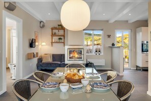 Ein Essbereich mit einem Glastisch, Korbstühlen und einem Brotkorb. Ein Kamin mit Feuer, eine Couch und ein Fenster mit Blick auf Grün sind ebenfalls vorhanden.