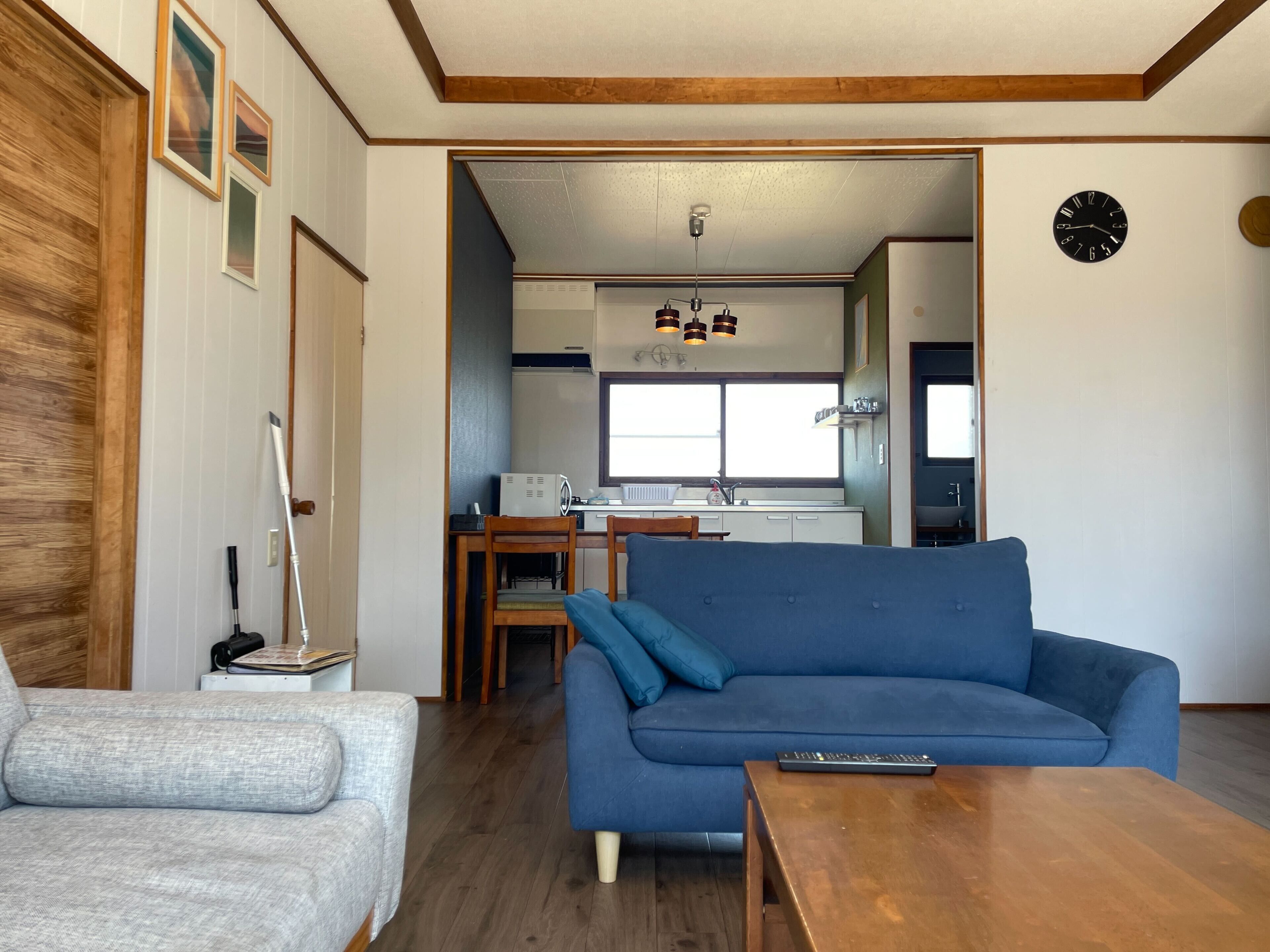 A living room with a blue sofa, a grey armchair, and a wooden coffee table. A kitchen is visible in the background with a window and a clock on the wall.