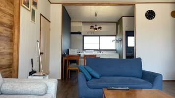 A living room with a blue sofa, a grey armchair, and a wooden coffee table. A kitchen is visible in the background with a window and a clock on the wall.