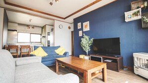 Una sala de estar con sofá, mesa de centro y televisor. Se ve una cocina al fondo.