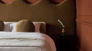 Ein ordentlich bezogenes Bett mit weißen Leinen und mehreren Kissen, eine runde Wandleuchte und ein kleiner Tisch mit einer Vase.