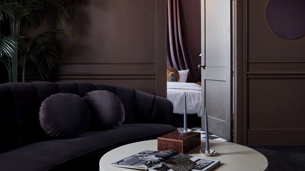 Una sala moderna con un sofá oscuro, una mesa de centro redonda con revistas, una planta en maceta y vistas a un dormitorio con cama y cortinas.