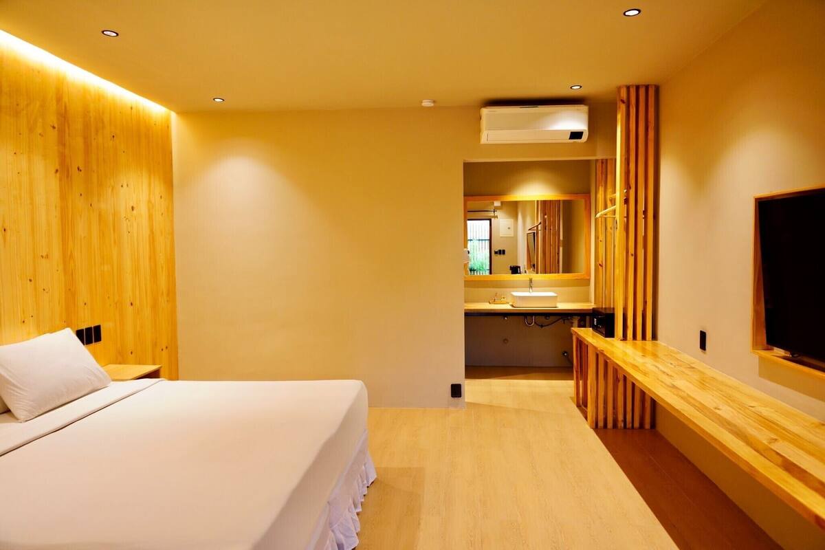 A modern hotel room with a wooden headboard, a large bed, a wooden bench, a TV, and a bathroom visible through an open door.