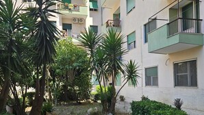 A multi-story residential building with balconies, a garden area with various plants, and a paved walkway.