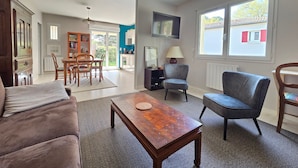 A living room with a sofa, armchair, coffee table, dining table, and chairs. There is a television mounted on the wall, a lamp, and a window with a view of the outside. - Cozy apartment with private garden, near beach, perfect for couples (Saint-Brevin-les-Pins)