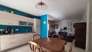 A dining area with a wooden table and chairs, a kitchen with white cabinets, and a blue accent wall. - Cozy apartment with private garden, near beach, perfect for couples (Saint-Brevin-les-Pins)