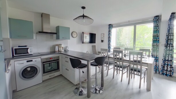 Een moderne keuken met een wasmachine, magnetron en fornuis. Er is een eethoek met een tafel en stoelen, en een groot raam met gordijnen.