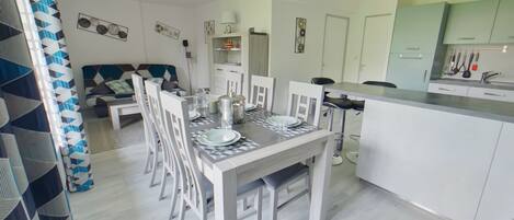 Une cuisine moderne avec des meubles blancs, un coin repas avec une table et des chaises blanches, et un salon avec un canapé et un rideau à motifs.