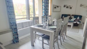 Een eetgedeelte met een witte tafel gedekt voor vier personen, een bank op de achtergrond en uitzicht op het landschap door grote ramen.