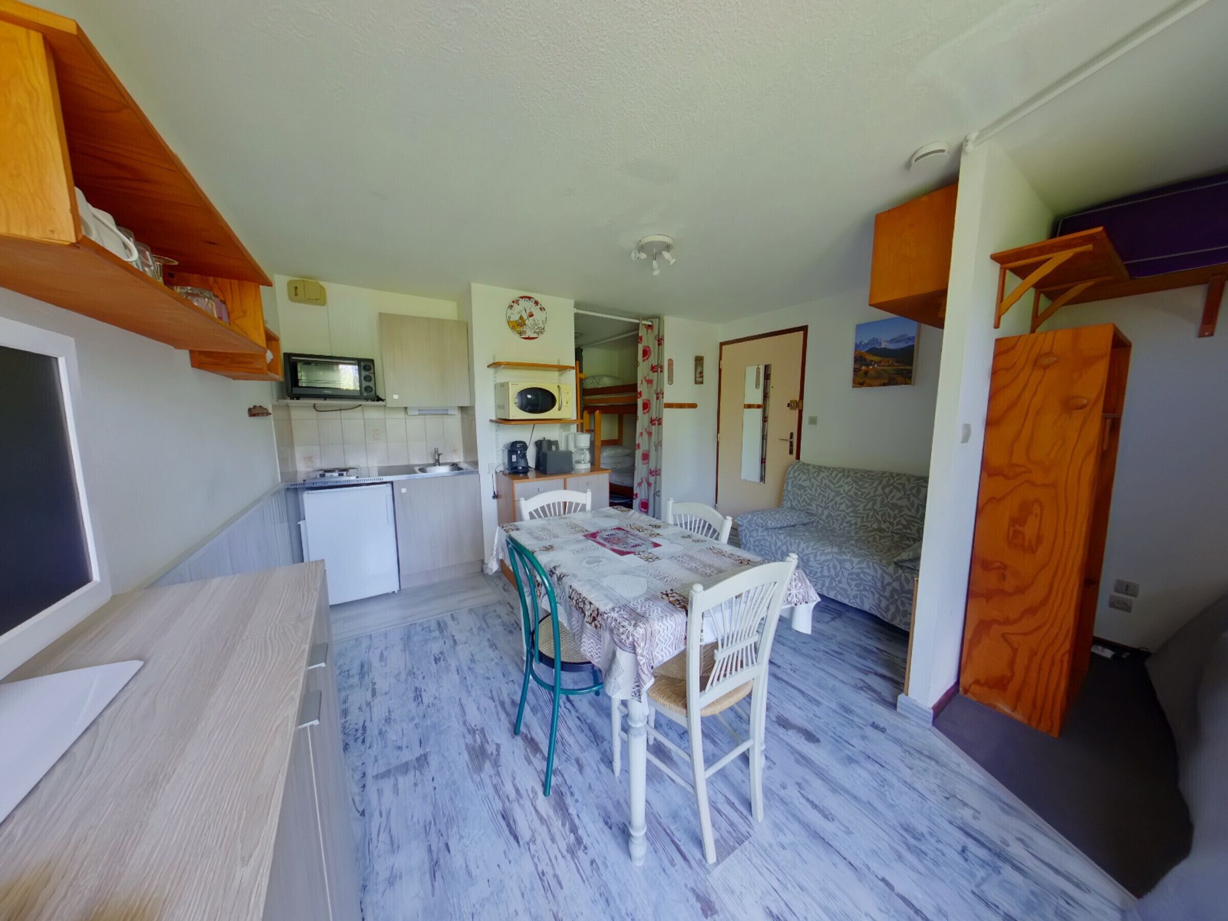 A compact kitchen with a microwave, sink, and small refrigerator. There is a dining table with chairs and a patterned tablecloth. A sofa is visible in the background.