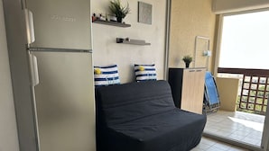 Ein gemütlicher Wohnraum mit Küchenzeile, inklusive Kühlschrank, einer Couch und einem kleinen Tisch. An der Wand befinden sich dekorative Kissen und ein Blumentopf.